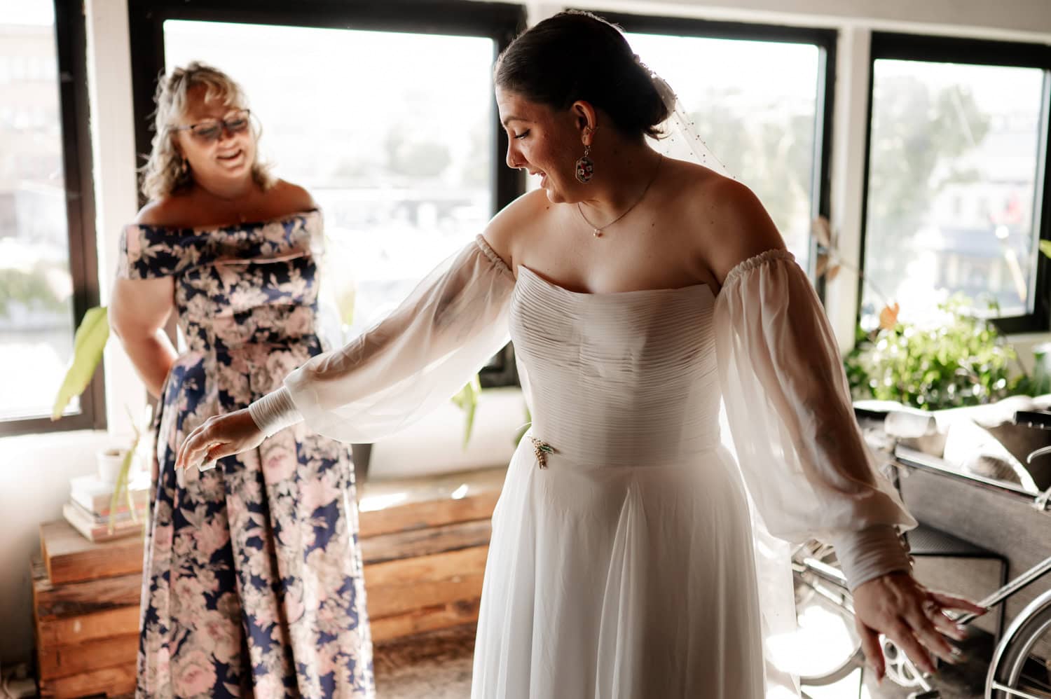 A woman in an off-shoulder wedding dress smiles brightly, standing in a sunlit room with large windows. Another woman, wearing a floral dress, looks at her with admiration. They are preparing for a wedding at Union/Pine.