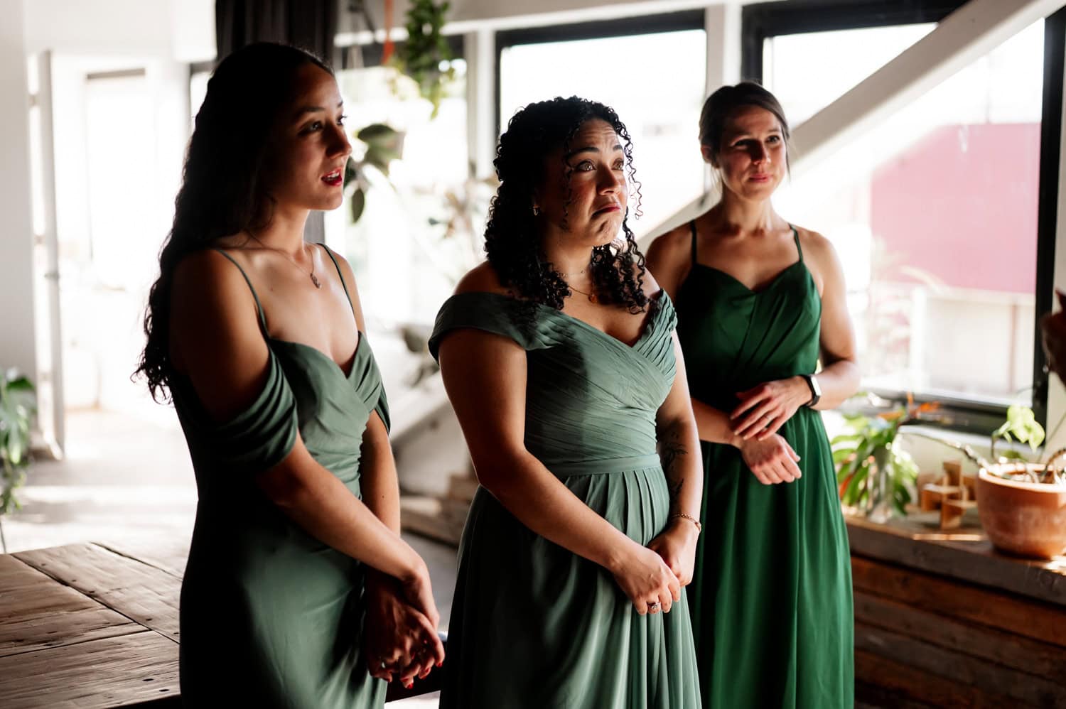 Three women in green dresses stand together at a wedding at Union/Pine, with sunlit plants surrounding them in the room.