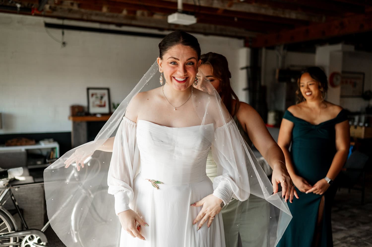 A bride in a white dress and veil smiles as her bridesmaids help her, with rustic decorations in the background at Union/Pine.