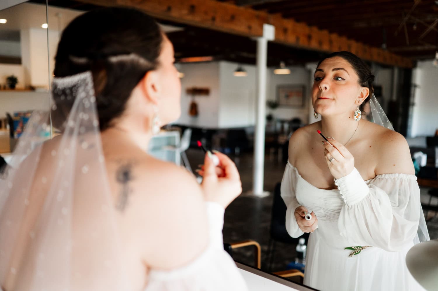 The bride, wearing an off-shoulder dress, applies lipstick as she looks at herself in the mirror. Behind her lies the enchanting setup of her wedding at Union/Pine.