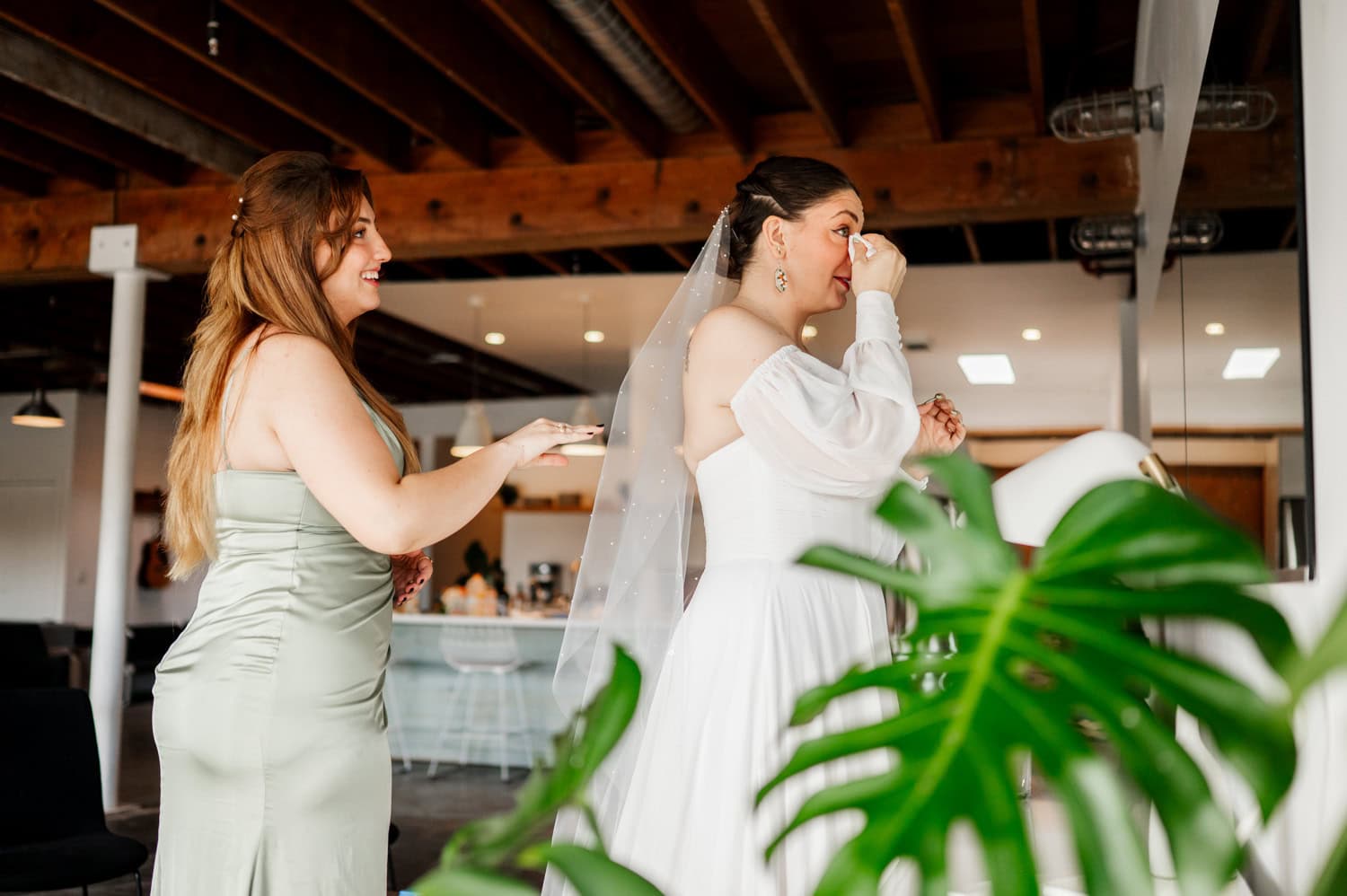 A bride wipes her tears with a tissue while a bridesmaid stands beside her, smiling supportively. They are inside Union/Pine, a venue featuring green plants and large wooden beams.