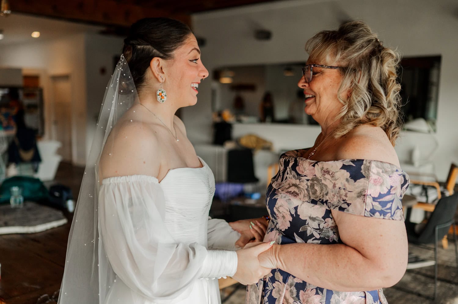 The bride in an elegant white dress holds hands and smiles with a woman wearing a vibrant floral dress inside Union/Pine.