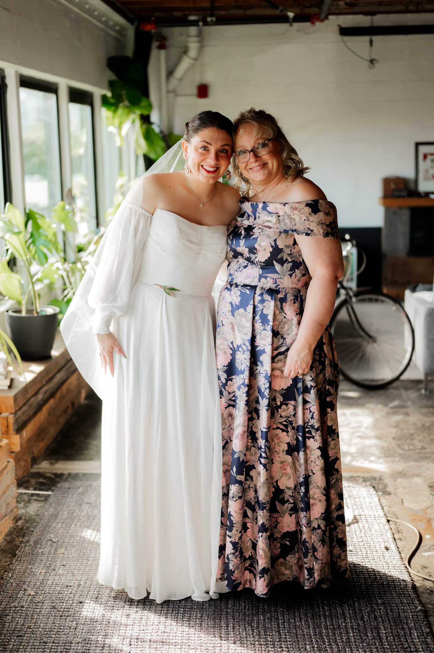 Two women in elegant dresses, one wearing white and the other in floral, pose together indoors by a window, smiling and capturing the joyful moment at a wedding held at Union/Pine.