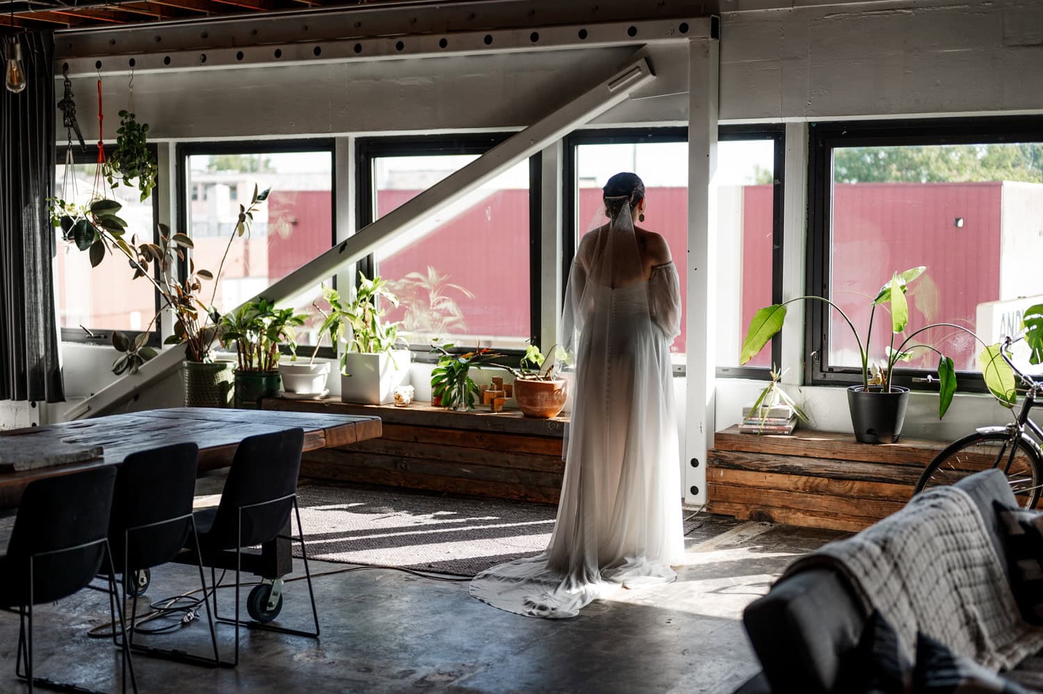 A bride in a long, sheer gown stands by large windows with plants around her in the industrial-chic setting of Union/Pine.