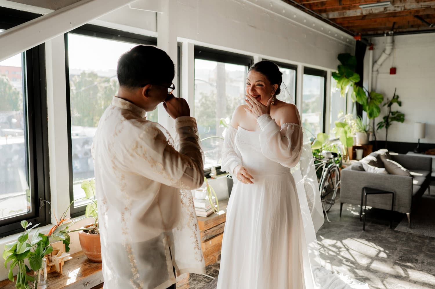 A couple in formal wedding attire shares a joyful moment near large windows and potted plants in a bright room at Union/Pine.