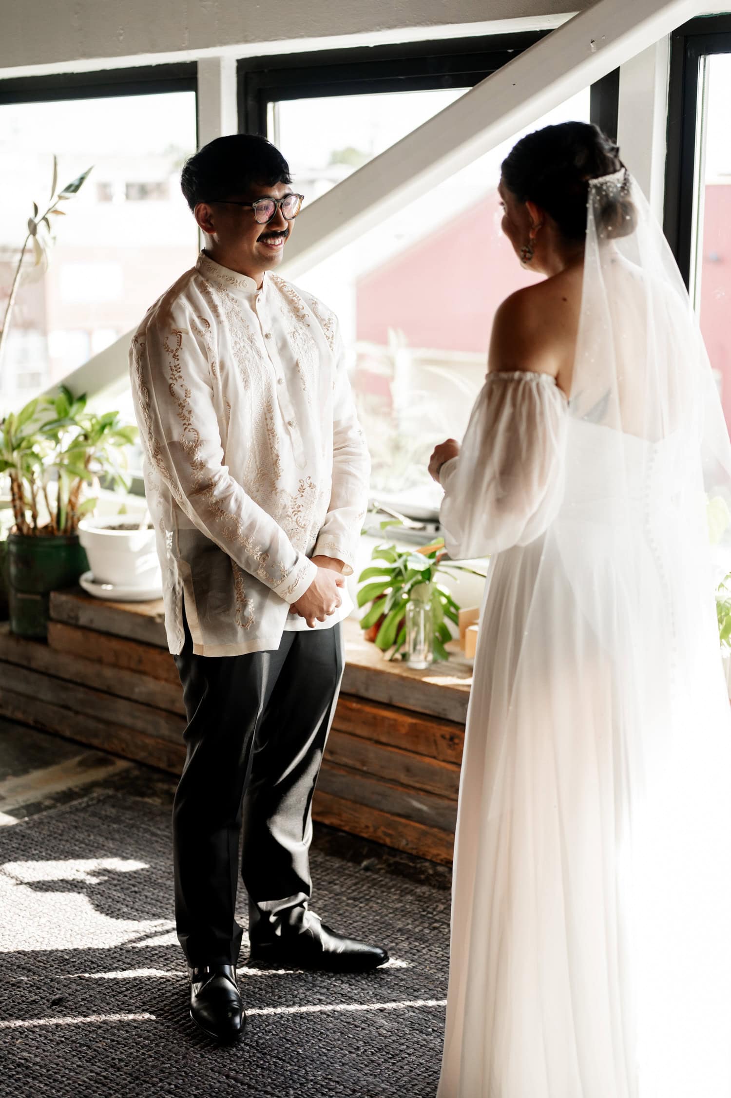 In a sunlit room at Union/Pine, the bride and groom exchange loving smiles. The groom wears a patterned shirt, and the bride is in a white dress, capturing an unforgettable wedding moment.