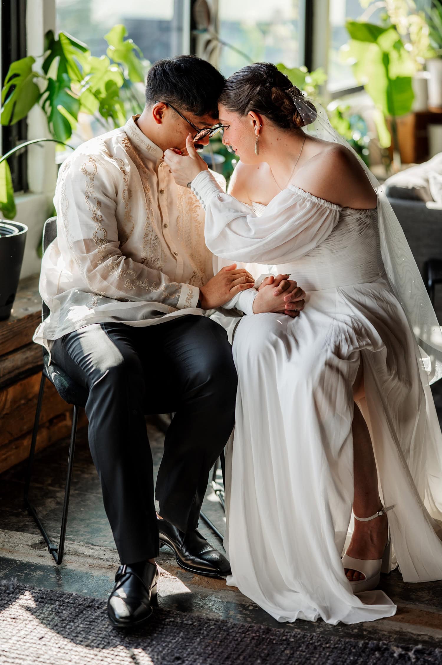 A couple in wedding attire sits together holding hands, sharing a tender moment near a sunlit window with plants, capturing the romance of their wedding at Union/Pine.