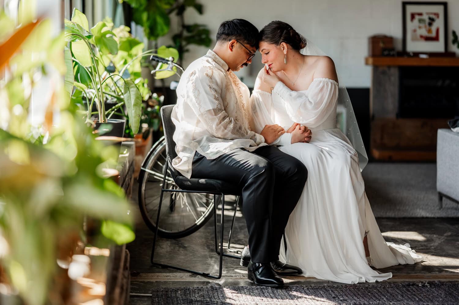 The bride and groom sit together holding hands, surrounded by lush plants with sunlight streaming through the window at their Union/Pine wedding.