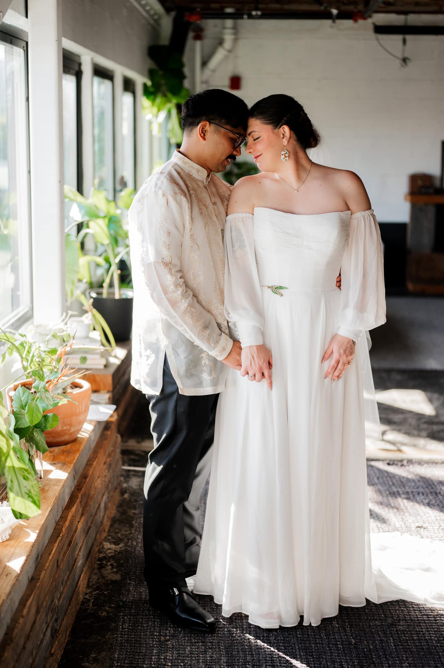 A couple in formal attire shares a tender moment at their wedding, standing by sunlit windows with lush green plants at Union/Pine.