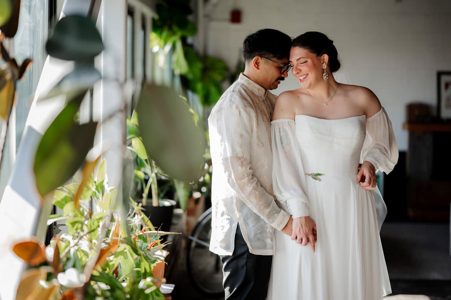 Bride and groom smiling and holding hands near large windows with indoor plants at their wedding at Union/Pine.