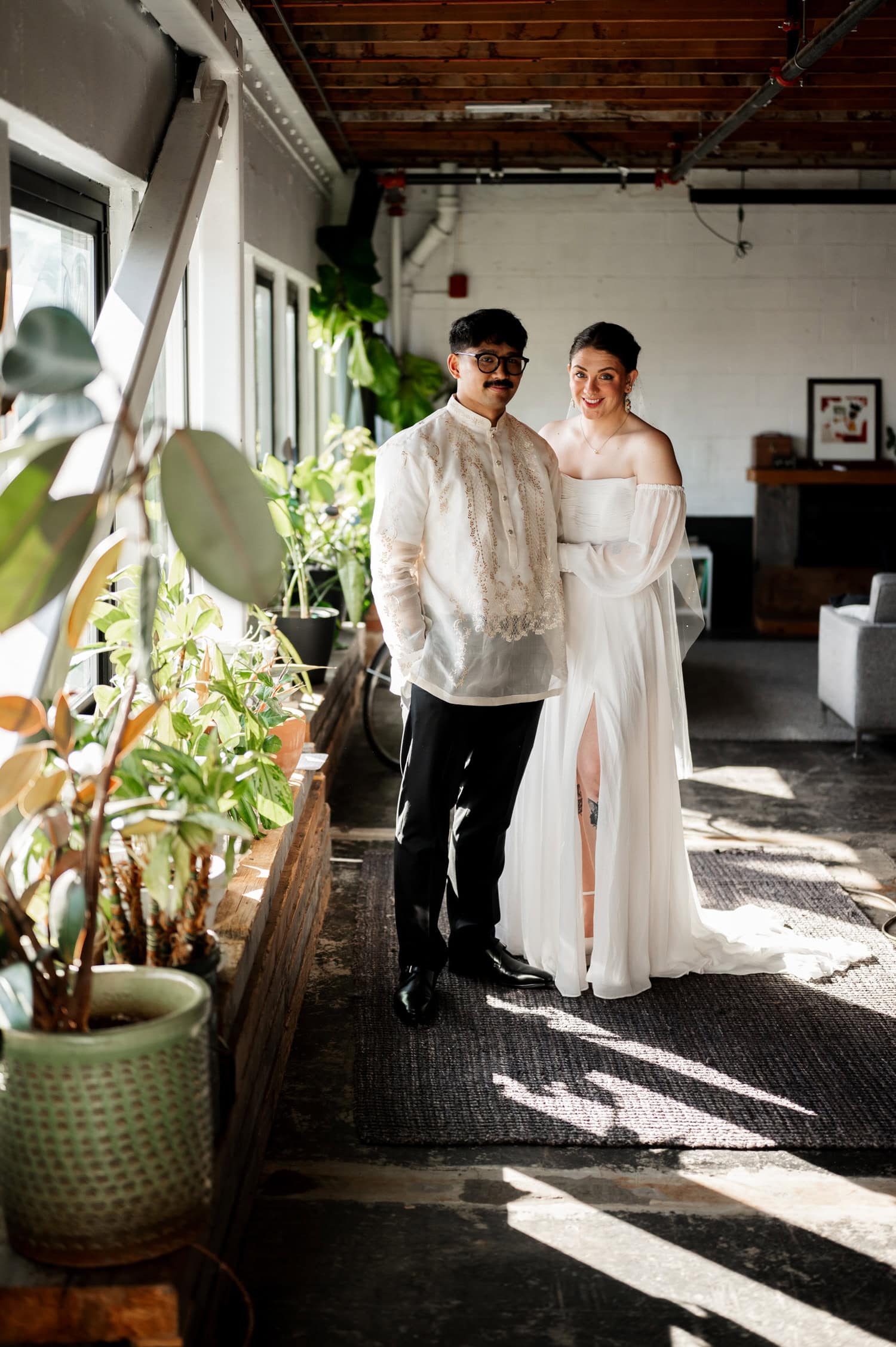 Sunlight streams through the large windows, illuminating a couple in wedding attire standing amidst potted plants at their Union/Pine wedding.