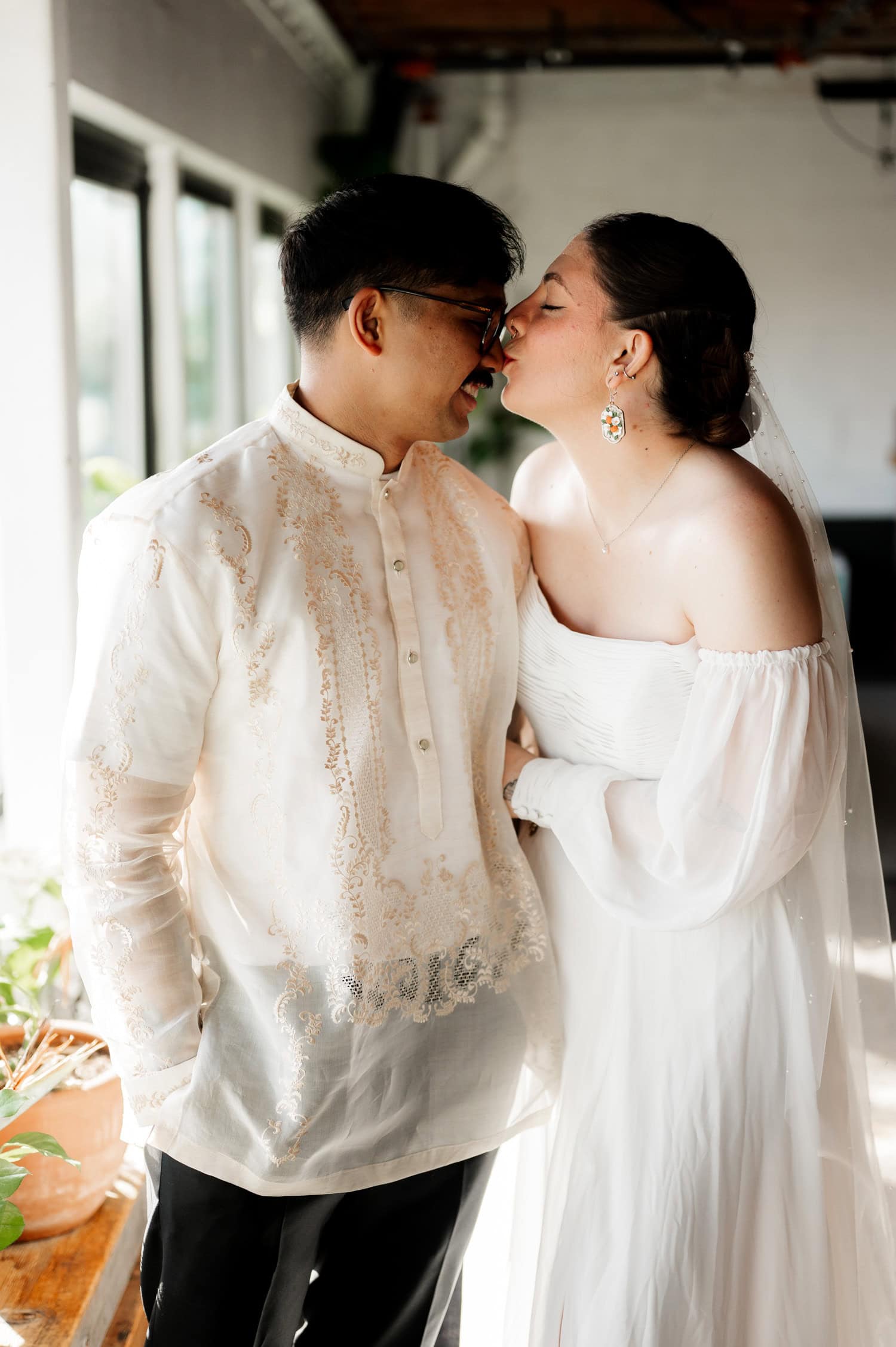 A couple in traditional attire stands close together indoors at their Union/Pine wedding, sharing a joyful moment with sunlight streaming through the window.
