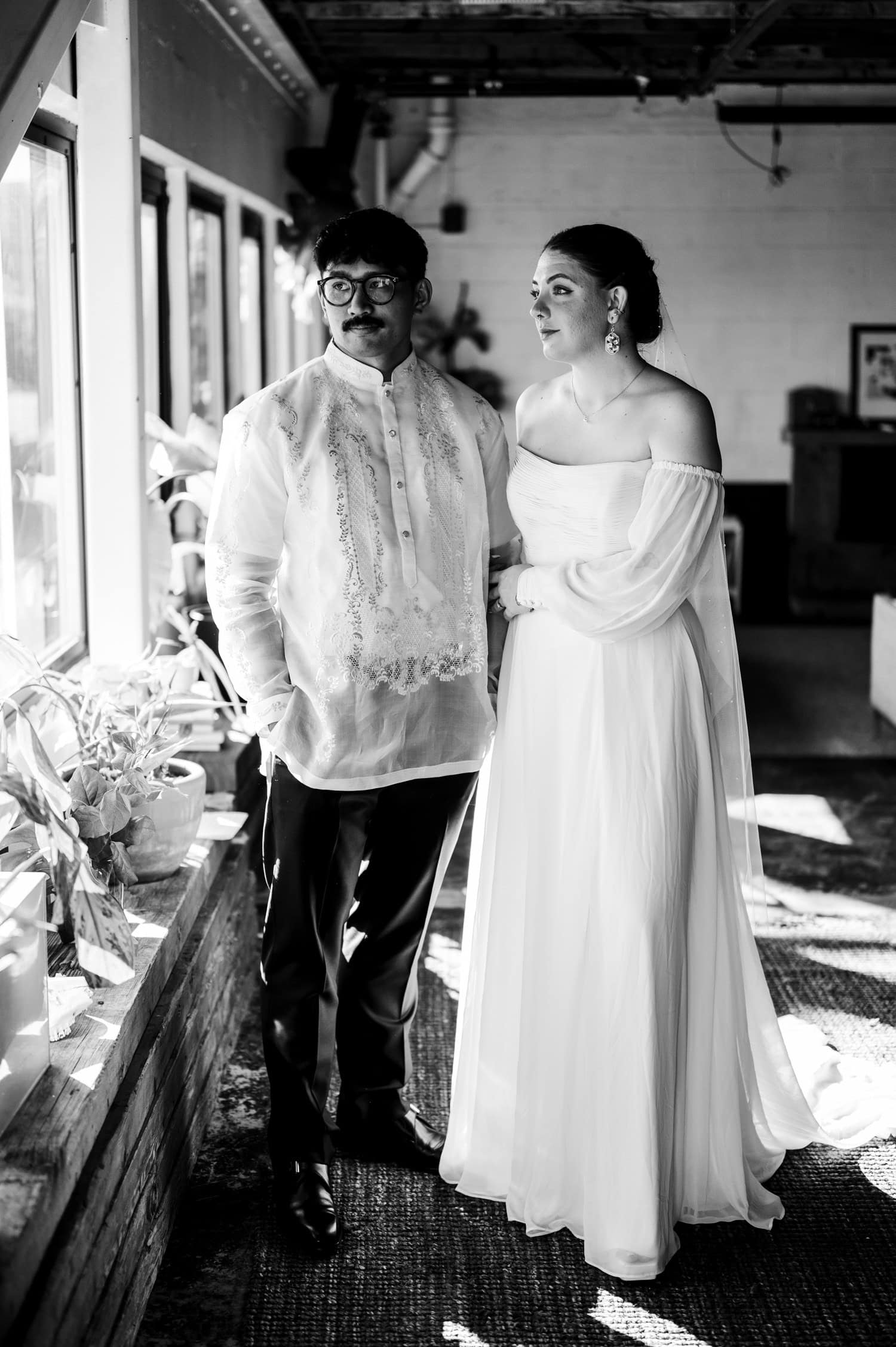 A couple stands by a window at their wedding at Union/Pine; the man wears a traditional barong, and the woman wears an elegant off-shoulder dress.