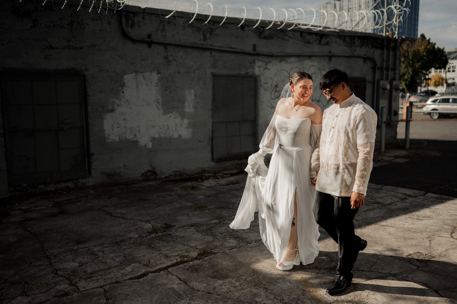 A couple in wedding attire holds hands and smiles as they walk near an industrial building, enjoying the sunlight after their wedding at Union/Pine.