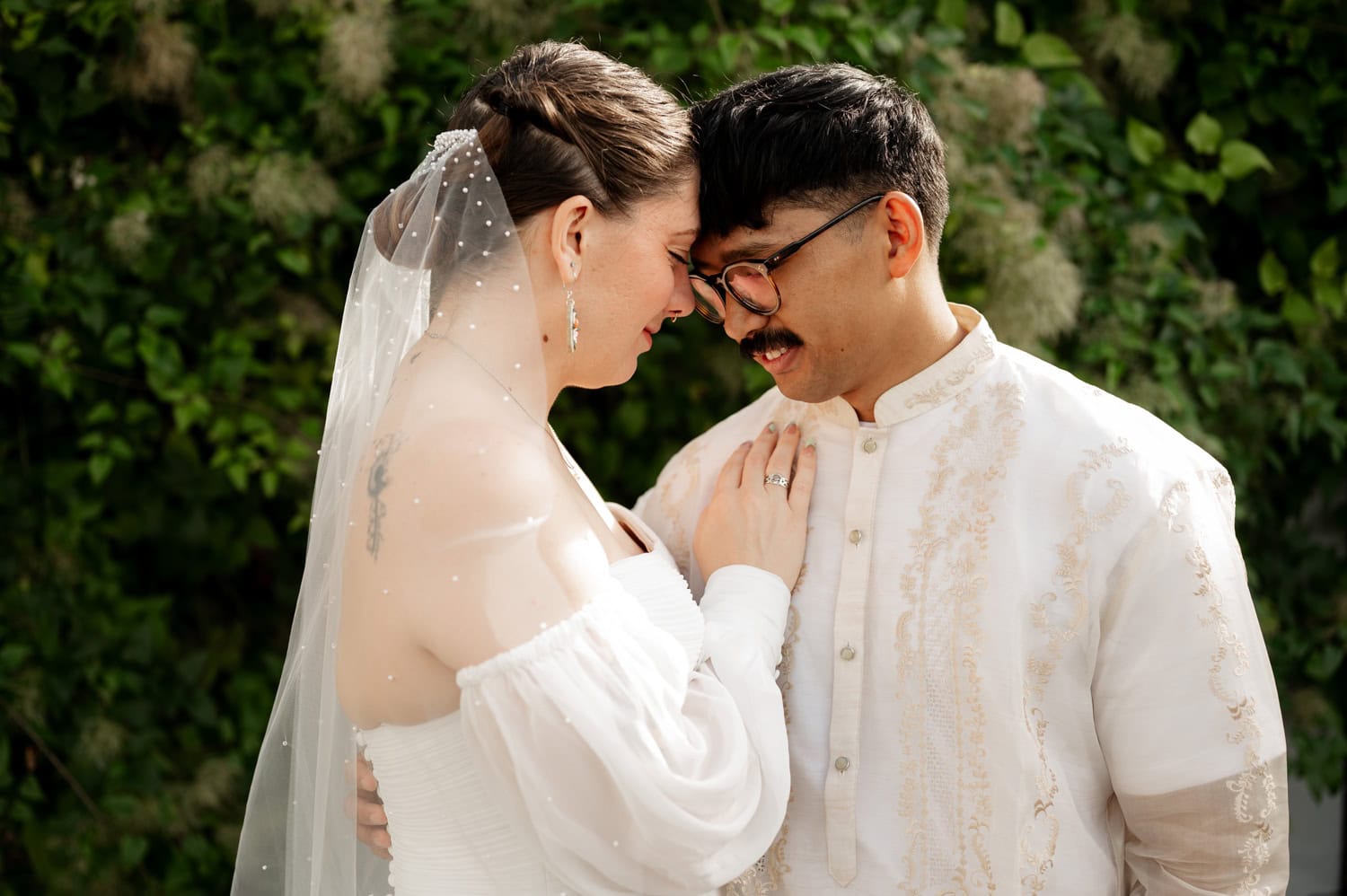 A couple in wedding attire shares a tender moment against a backdrop of lush, green foliage at their Union/Pine wedding.