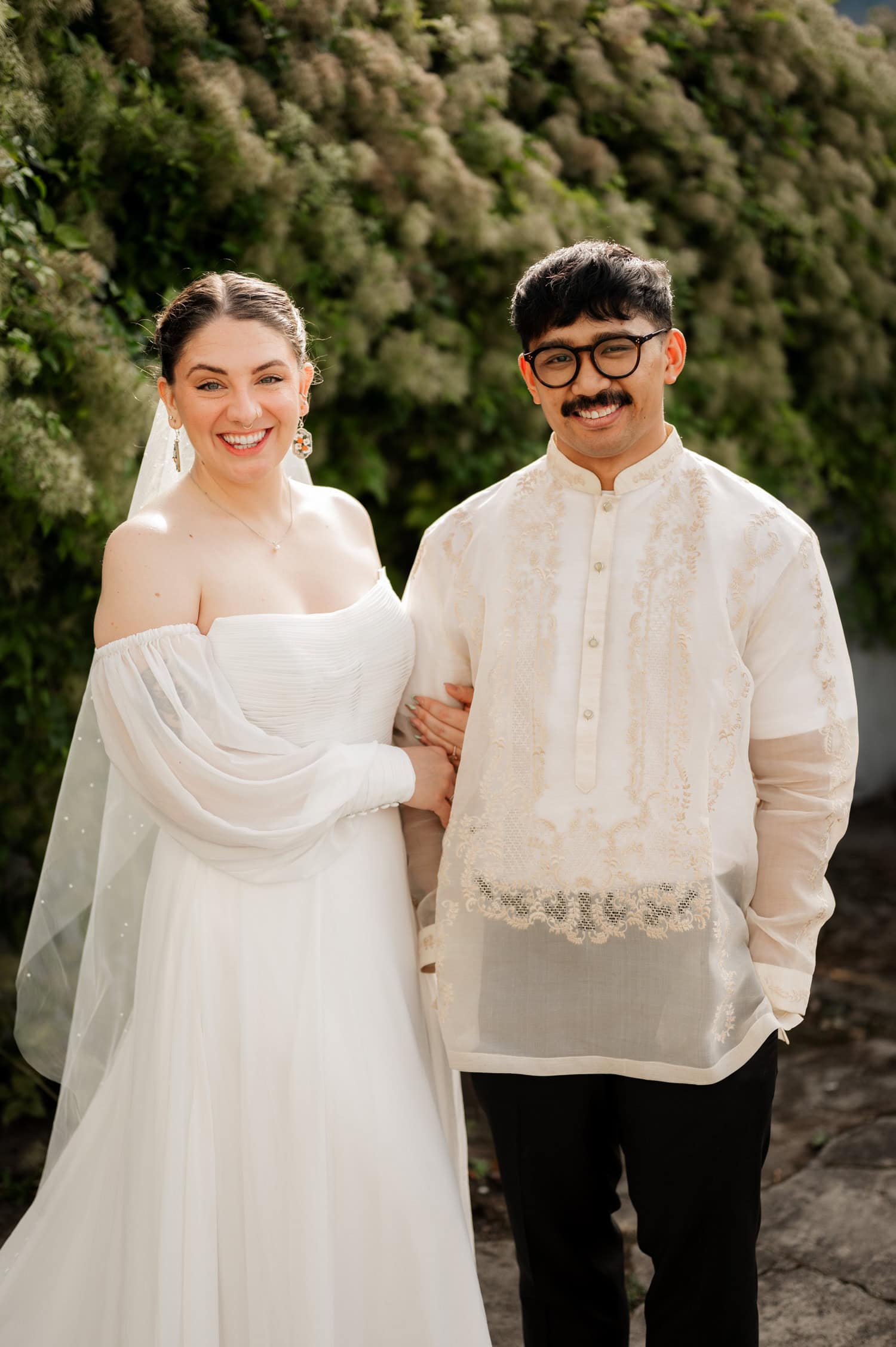 A bride in a white gown smiles happily next to the groom in traditional attire. They are surrounded by vibrant greenery at their wedding venue, Union/Pine.