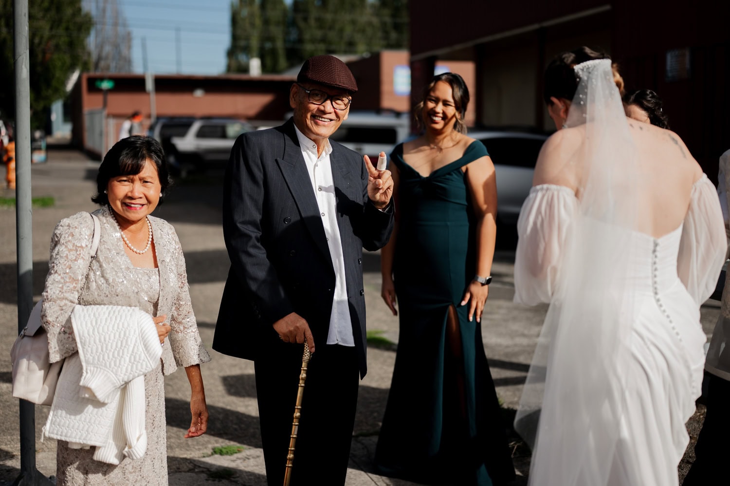 A joyful group gathers outdoors at a wedding at Union/Pine; an older man makes a peace sign while surrounded by women in dresses, including the bride in her gown.
