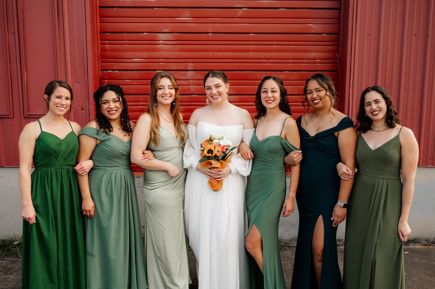 A bride in a white dress smiles with her bridesmaids dressed in green in front of a red garage door at Union/Pine.