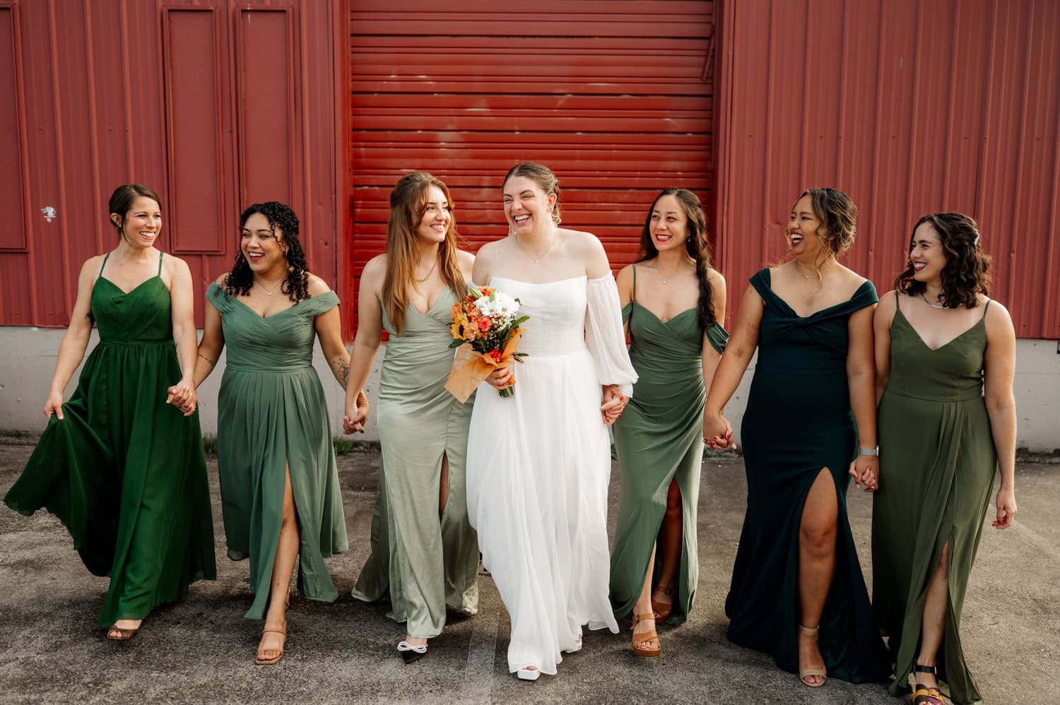 Bride in a white dress stands with bridesmaids wearing green dresses, all smiling and holding hands. They are positioned against a red wall at a wedding held at Union/Pine.