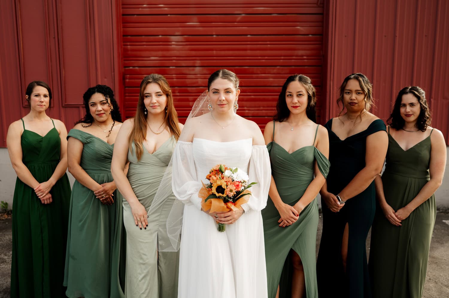 In front of a striking red backdrop, the bride in a white dress holds a bouquet and stands with six bridesmaids wearing green dresses at Union/Pine.