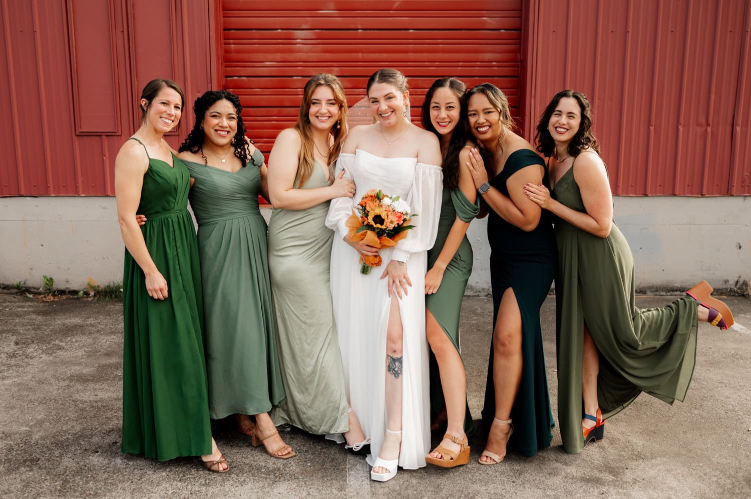 The bride, wearing a white gown and holding a bouquet, stands with six bridesmaids in green dresses. They are posed in front of a vivid red wall at Union/Pine for their wedding.