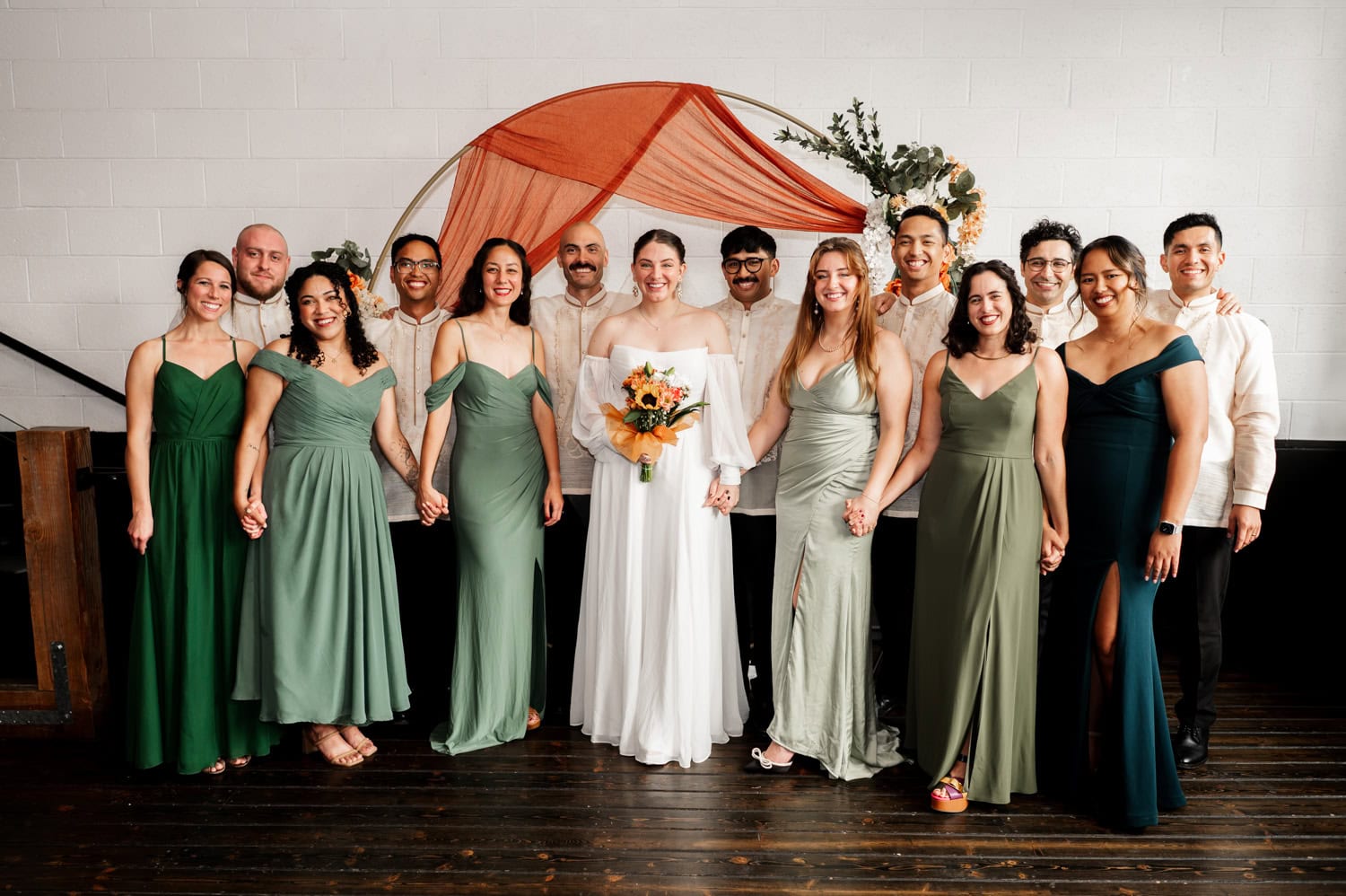 The bride holds a bouquet while standing with her wedding party, who are wearing coordinated green dresses. They are positioned in front of an orange circular backdrop at a wedding at Union/Pine.
