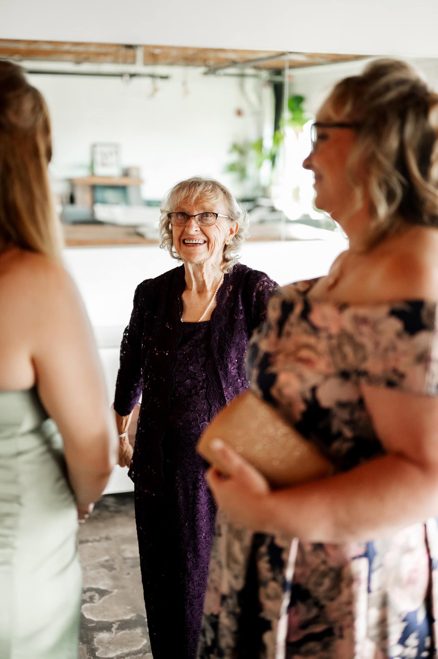Three women smiling and chatting indoors at a wedding, one elderly woman wearing a purple dress and two middle-aged women in floral dresses.