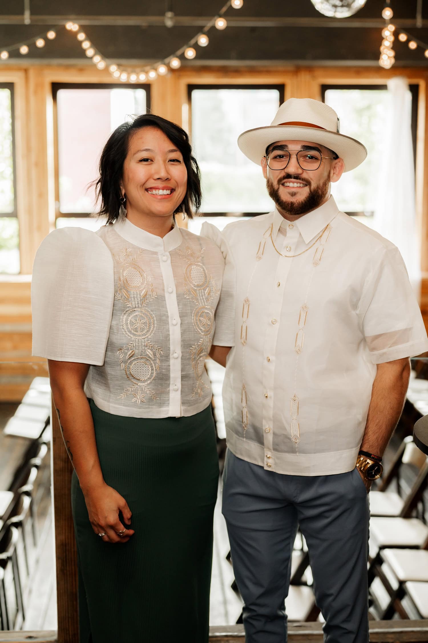 A smiling couple poses indoors under string lights, both wearing white shirts, capturing a special moment from their wedding at Union/Pine.