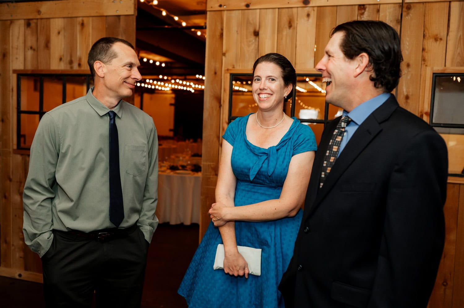 Three people in formal attire smile and chat in a warmly lit room with wooden walls and string lights, capturing the joyous ambiance of a wedding at Union/Pine.