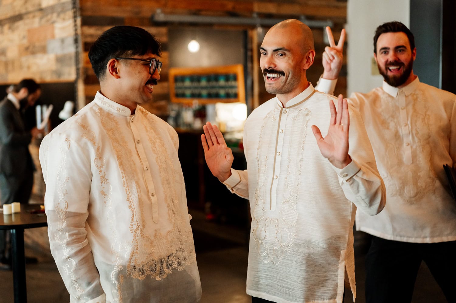 Three men in traditional embroidered shirts laughing together at a wedding. One man playfully holds up a peace sign behind anothers head, enhancing the joyful mood.