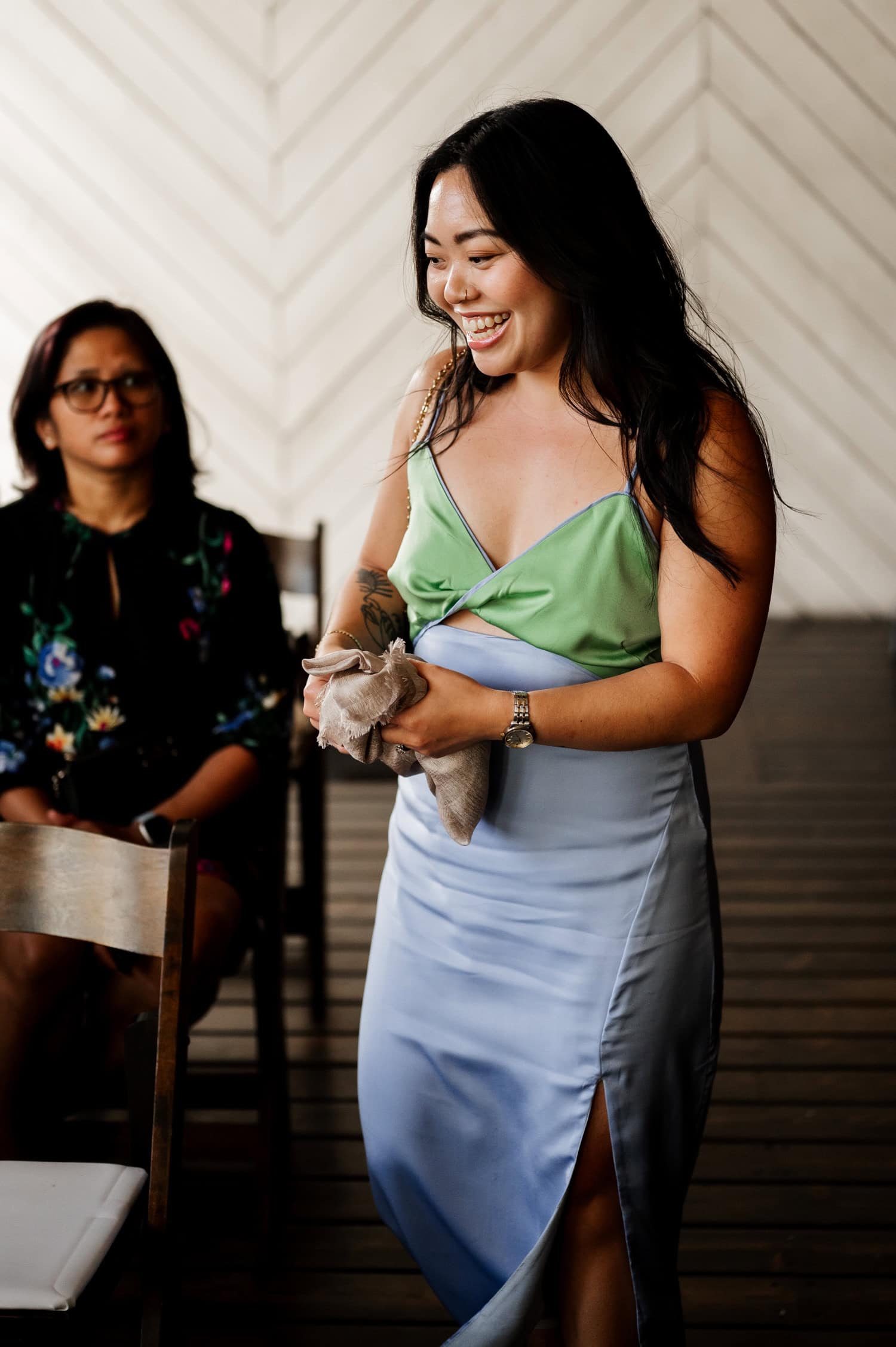 A smiling woman in a green and blue dress holds a napkin at the wedding at Union/Pine. Another woman sits on a wooden chair nearby.