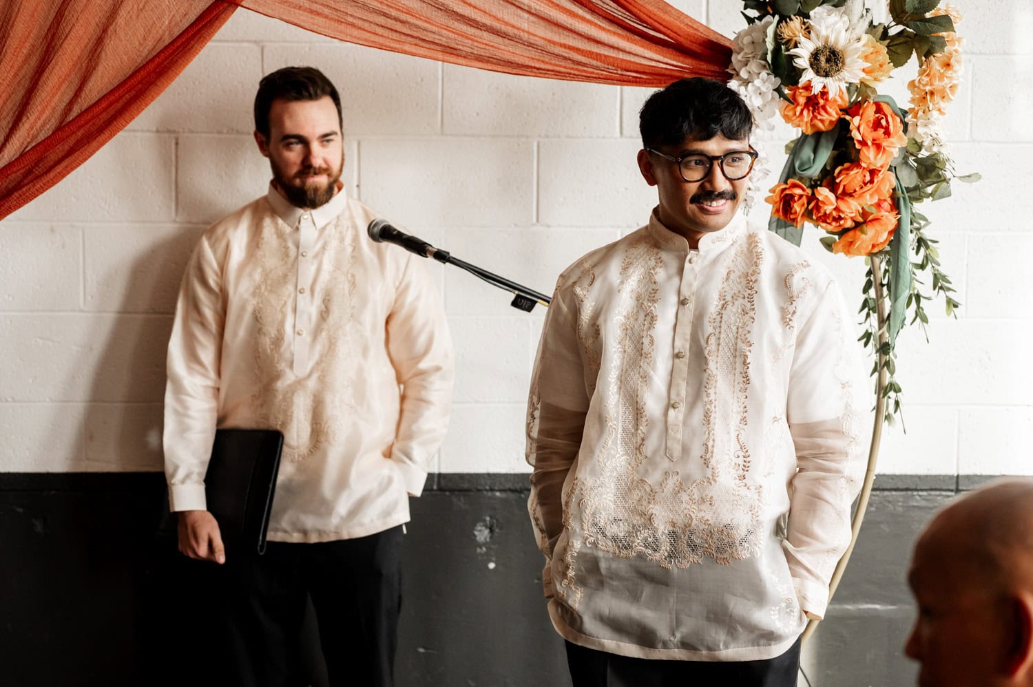 Two men in Barong Tagalog stand smiling by a microphone and floral arrangement at Union/Pine during a wedding.