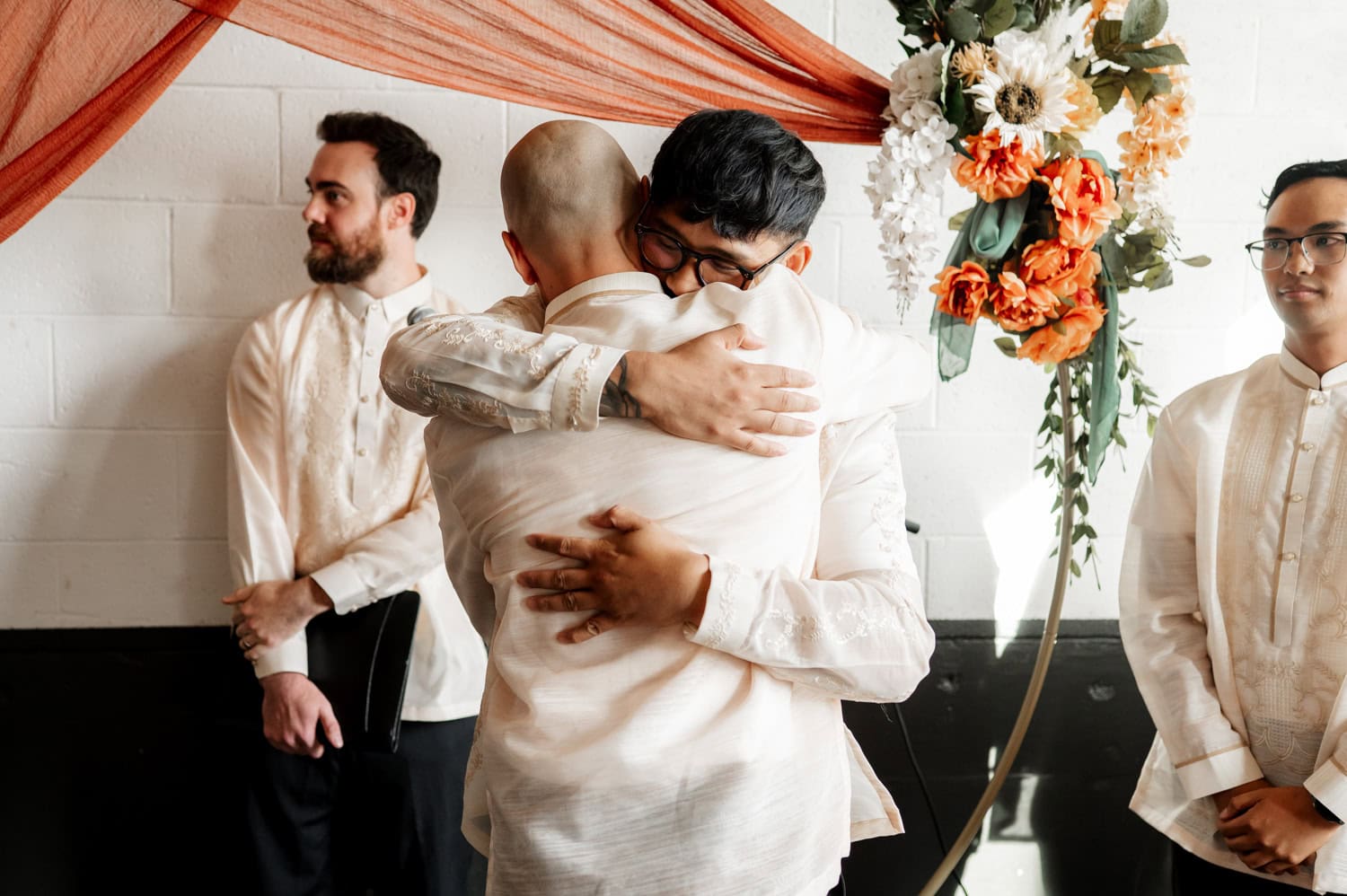 Two people in traditional attire embrace warmly at a wedding at Union/Pine, surrounded by floral decorations and onlookers.