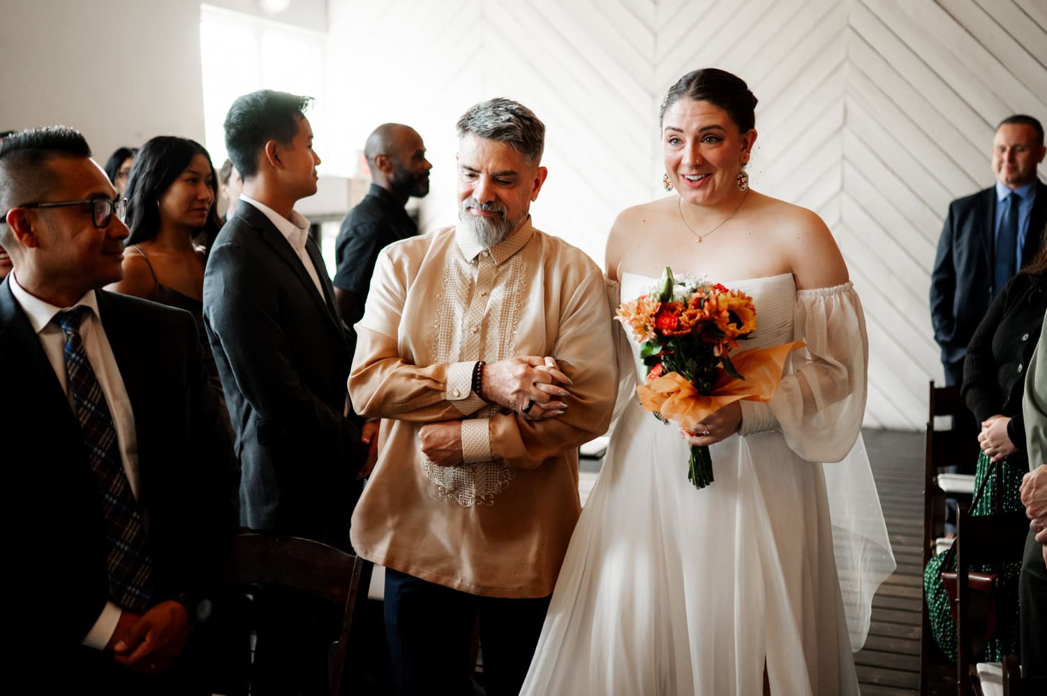 A bride in a white dress smiles while holding sunflowers, being escorted down the aisle by a man in a traditional embroidered shirt during their wedding at Union/Pine.