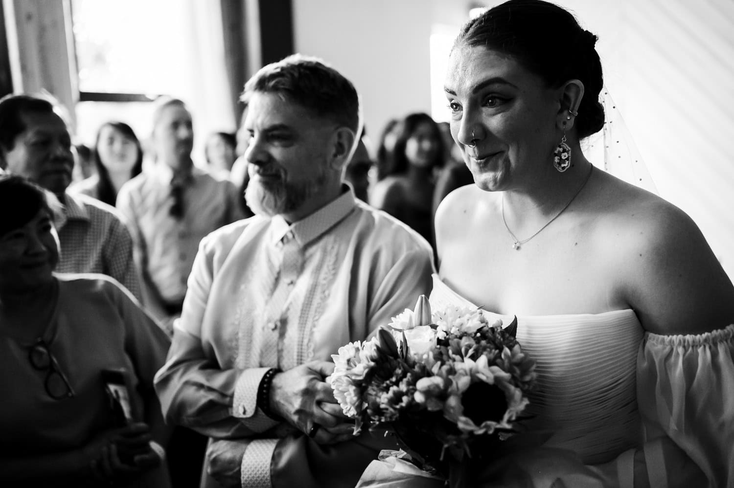 A bride smiles while holding her bouquet as she walks down the aisle with a man beside her at Union/Pine. People watch the procession in the background in this black and white photo.
