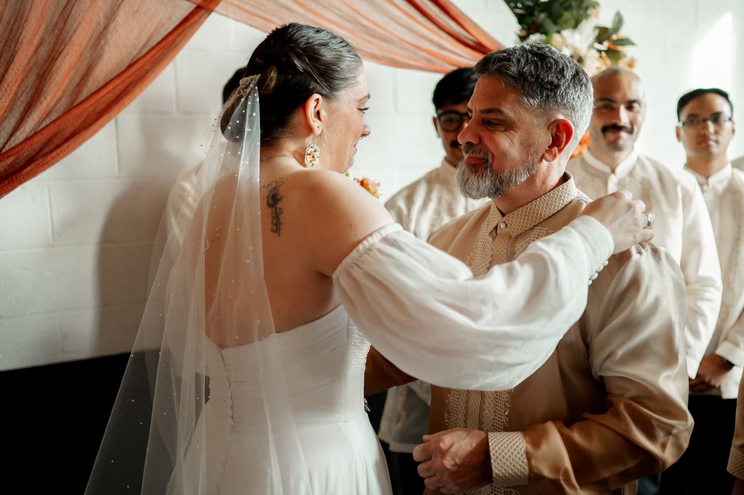 The bride and groom smile at each other surrounded by groomsmen in a warmly lit room with draped fabric, creating a beautiful wedding scene at Union/Pine.