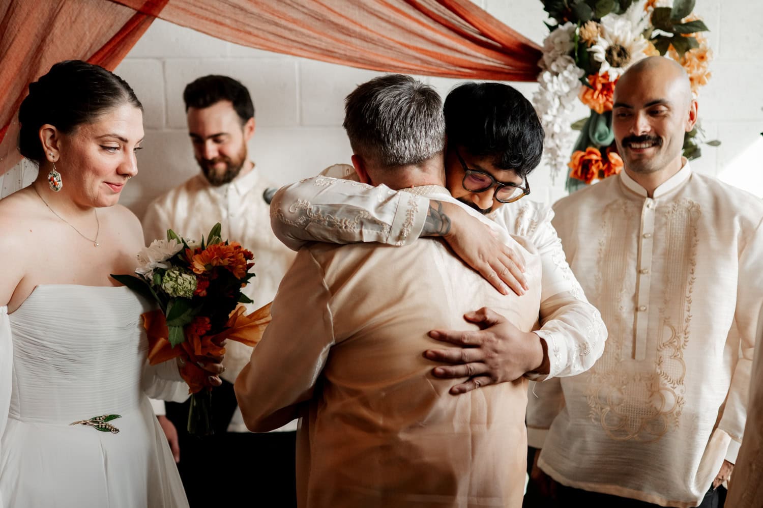 At a vibrant wedding at Union/Pine, the bride holds a bouquet of flowers and stands amidst three men wearing elegant barongs. Two of the men share a warm embrace, while everyone displays bright smiles, capturing a moment filled with joy and camaraderie.