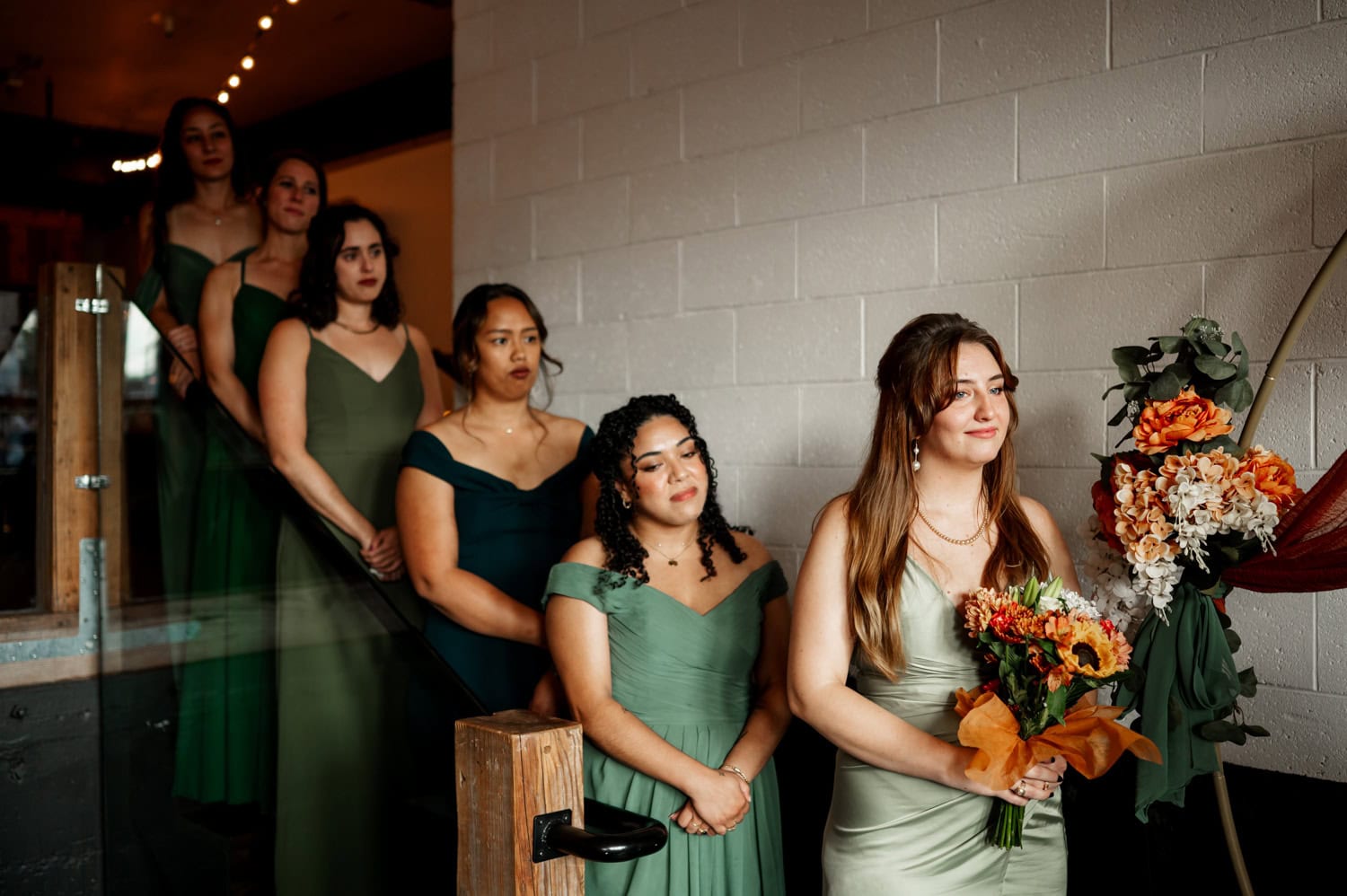 Bridesmaids in green dresses stand on steps next to a large flower arrangement, holding bouquets indoors at a wedding at Union/Pine.