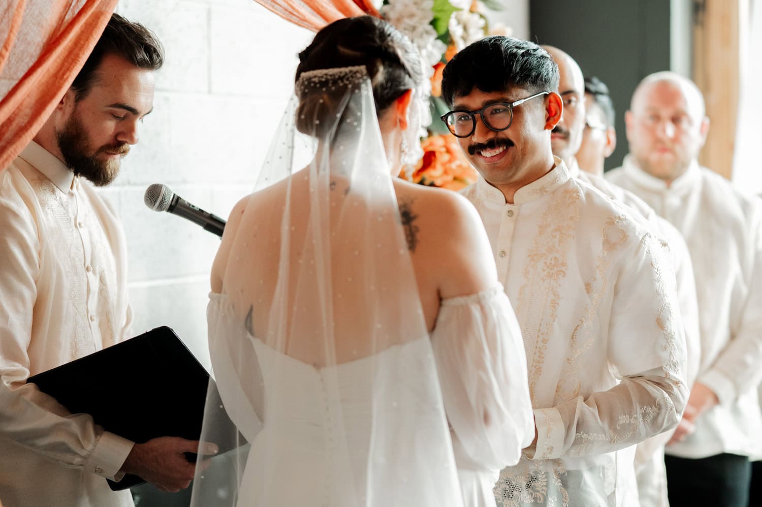 A bride and groom stand facing each other during their wedding ceremony at Union/Pine, with the officiant and groomsmen positioned nearby.