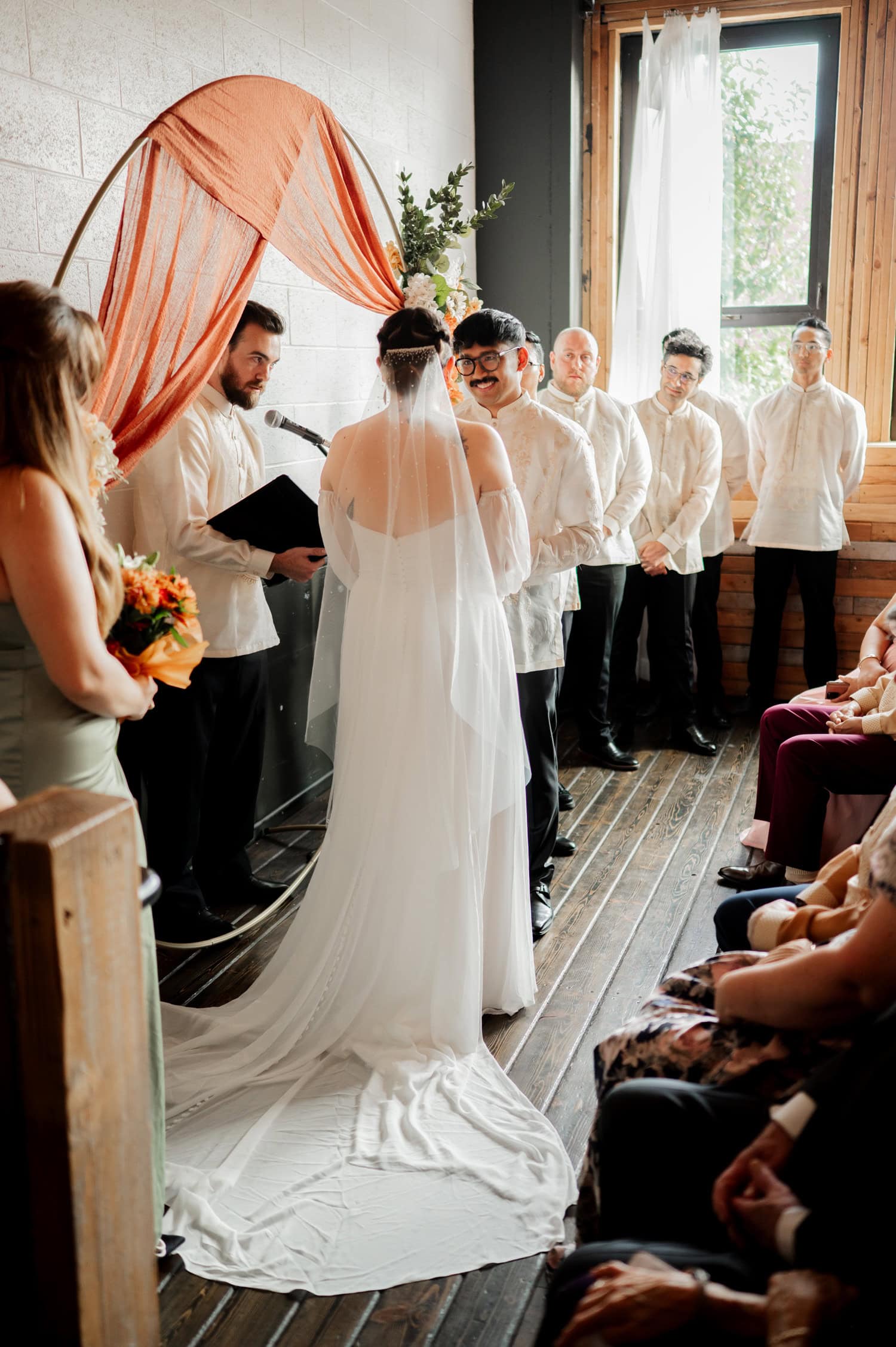 A bride and groom stand at the altar in a warmly lit room, surrounded by their wedding party and guests during their wedding ceremony at Union/Pine.