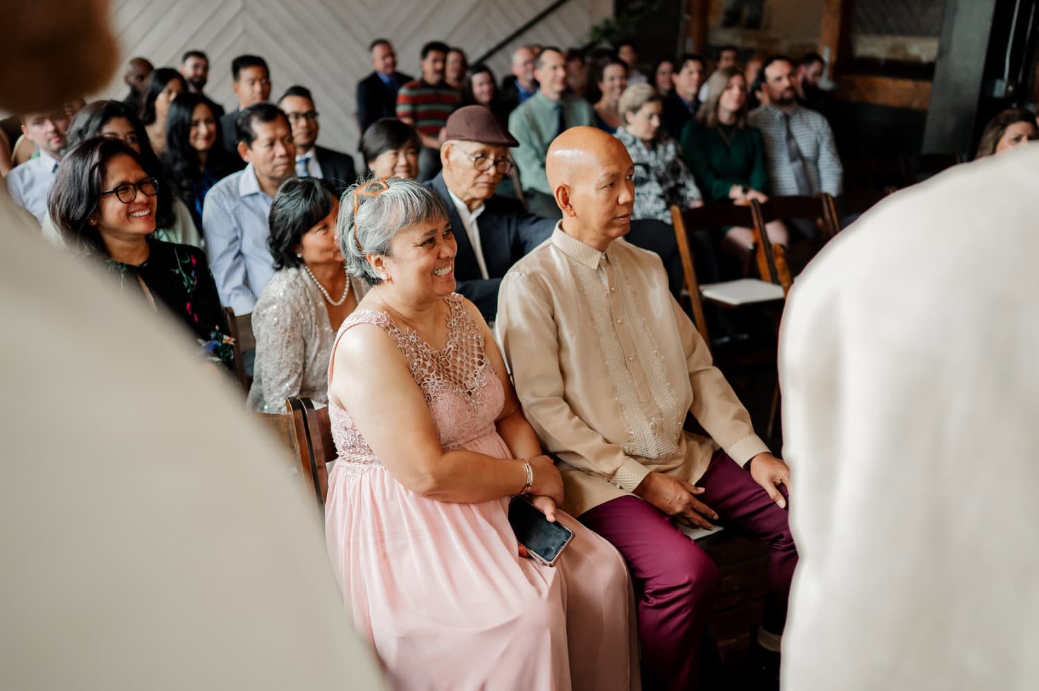A couple in elegant attire stands at the center of a wedding ceremony at Union/Pine, surrounded by smiling guests seated in rows. The indoor venue has a warm and formal ambiance, capturing moments of celebration and happiness.
