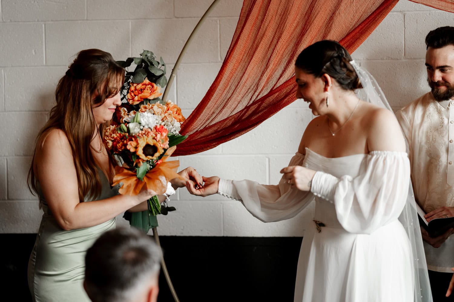 The bride, wearing a beautiful white dress, places a ring on the finger of another woman holding a bouquet, with fabric drapes creating a circular backdrop.