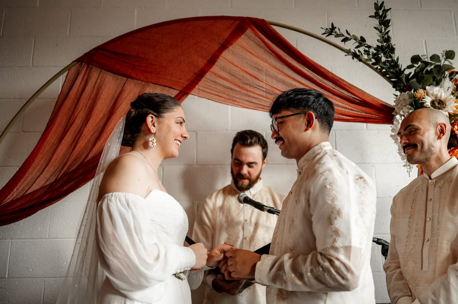 Bride and groom exchanging rings with joyful expressions, accompanied by two groomsmen and festive decorations in the background at Union/Pine wedding venue.