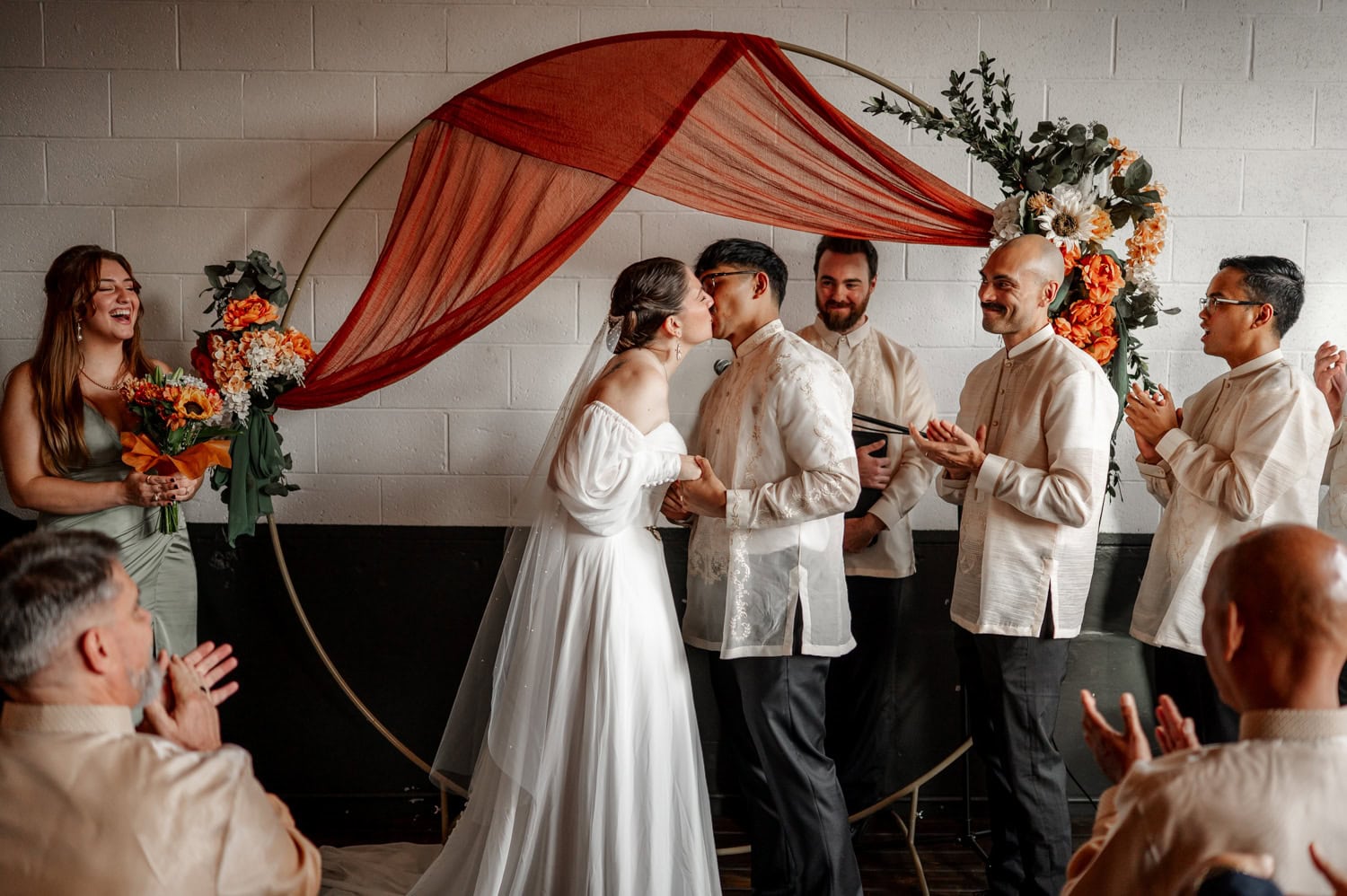 A couple kisses under a floral arch at their wedding, with guests smiling around them at Union/Pine.