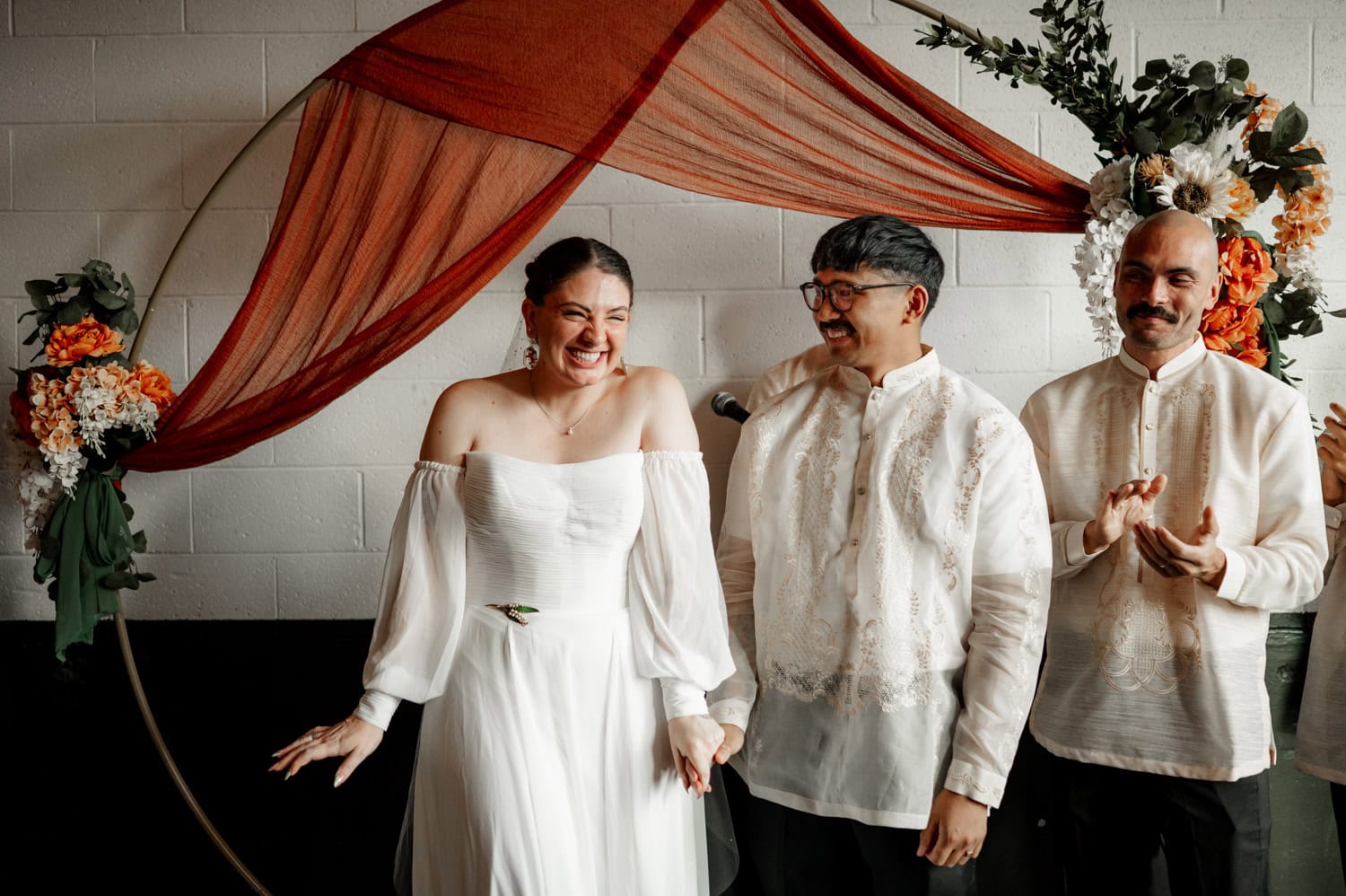The bride and groom smile brightly under a floral arch with a reddish-brown drape during their wedding at Union/Pine.