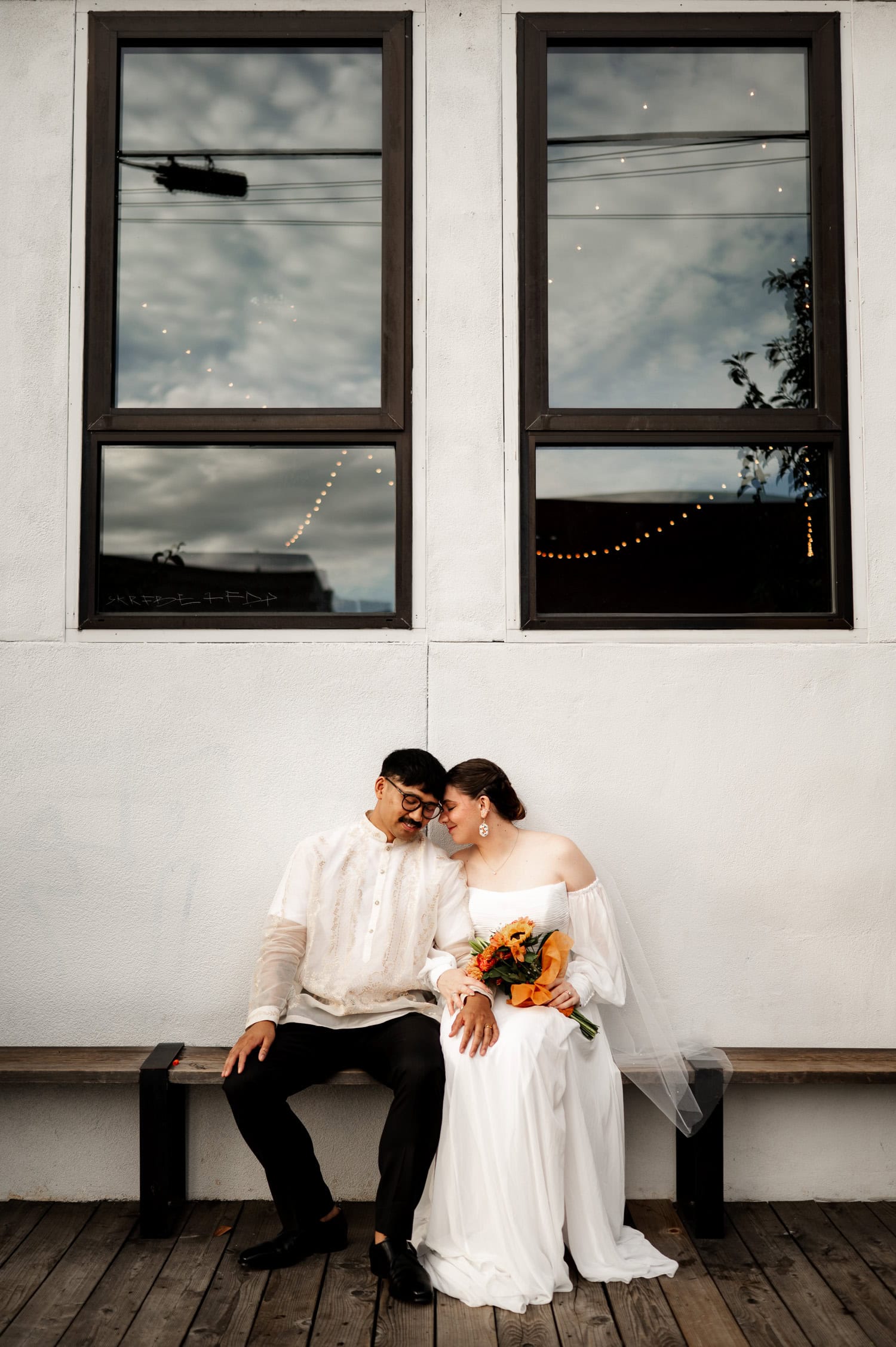 A bride and groom sit smiling on a bench in front of the large windows at Union/Pine.
