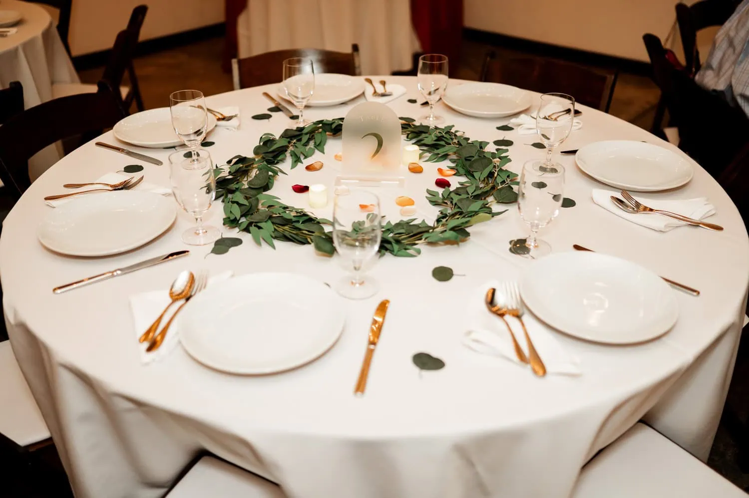 A round table with a white tablecloth is set for a wedding at Union/Pine, featuring plates, glassware, gold cutlery, and a leafy centerpiece.