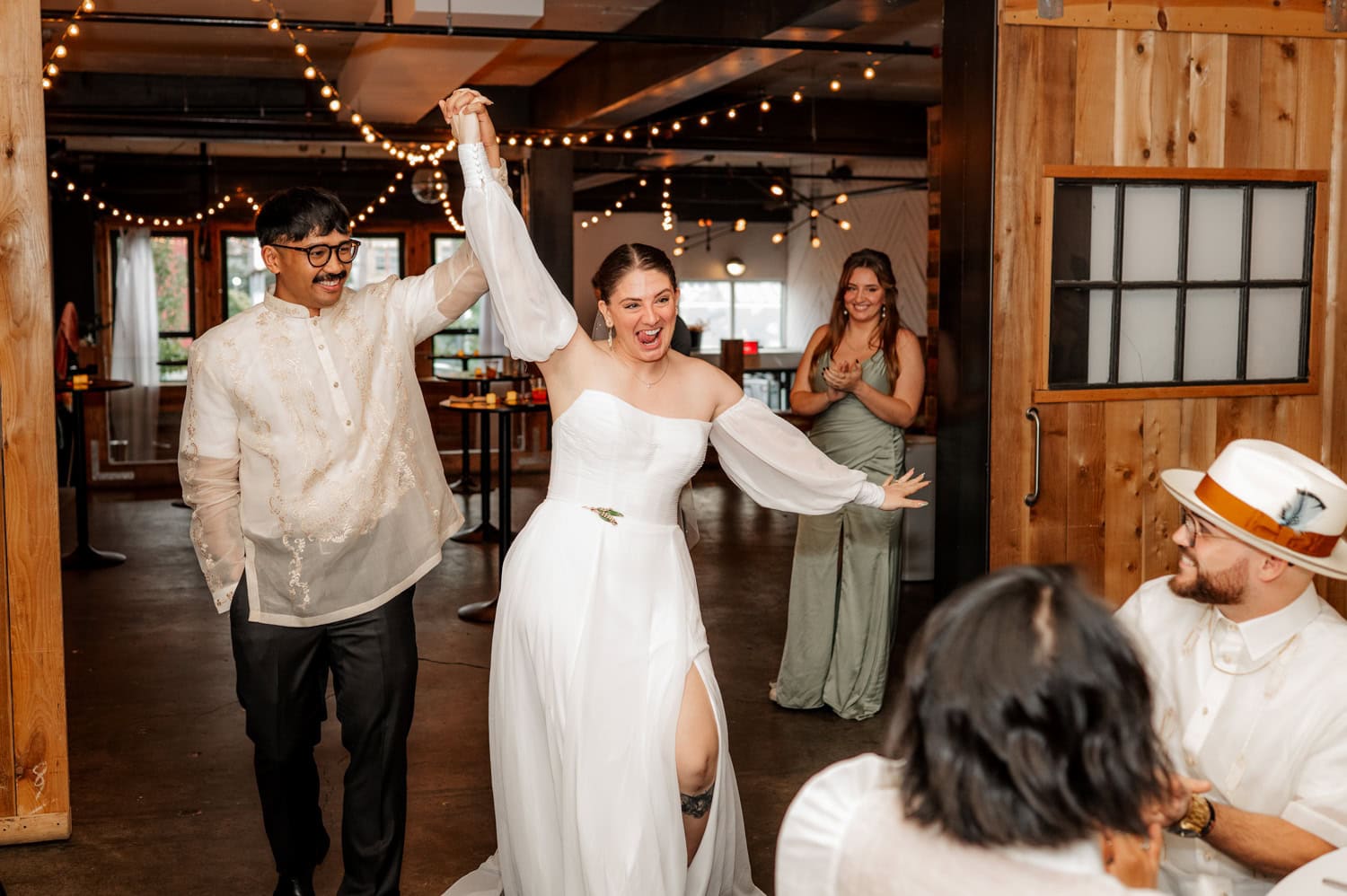 A joyful couple walks hand-in-hand into their wedding at Union/Pine, smiling as they are surrounded by guests. The venue is warmly lit, creating an inviting atmosphere.