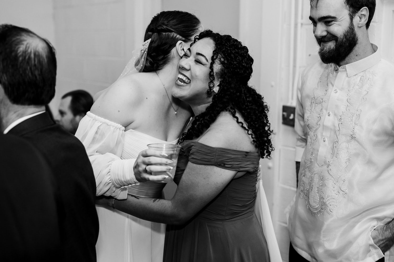 A bride in a formal dress hugs a smiling woman with curly hair, both wearing elegant attire, at a wedding held at Union/Pine. A joyful man stands close to them, sharing in the celebration.