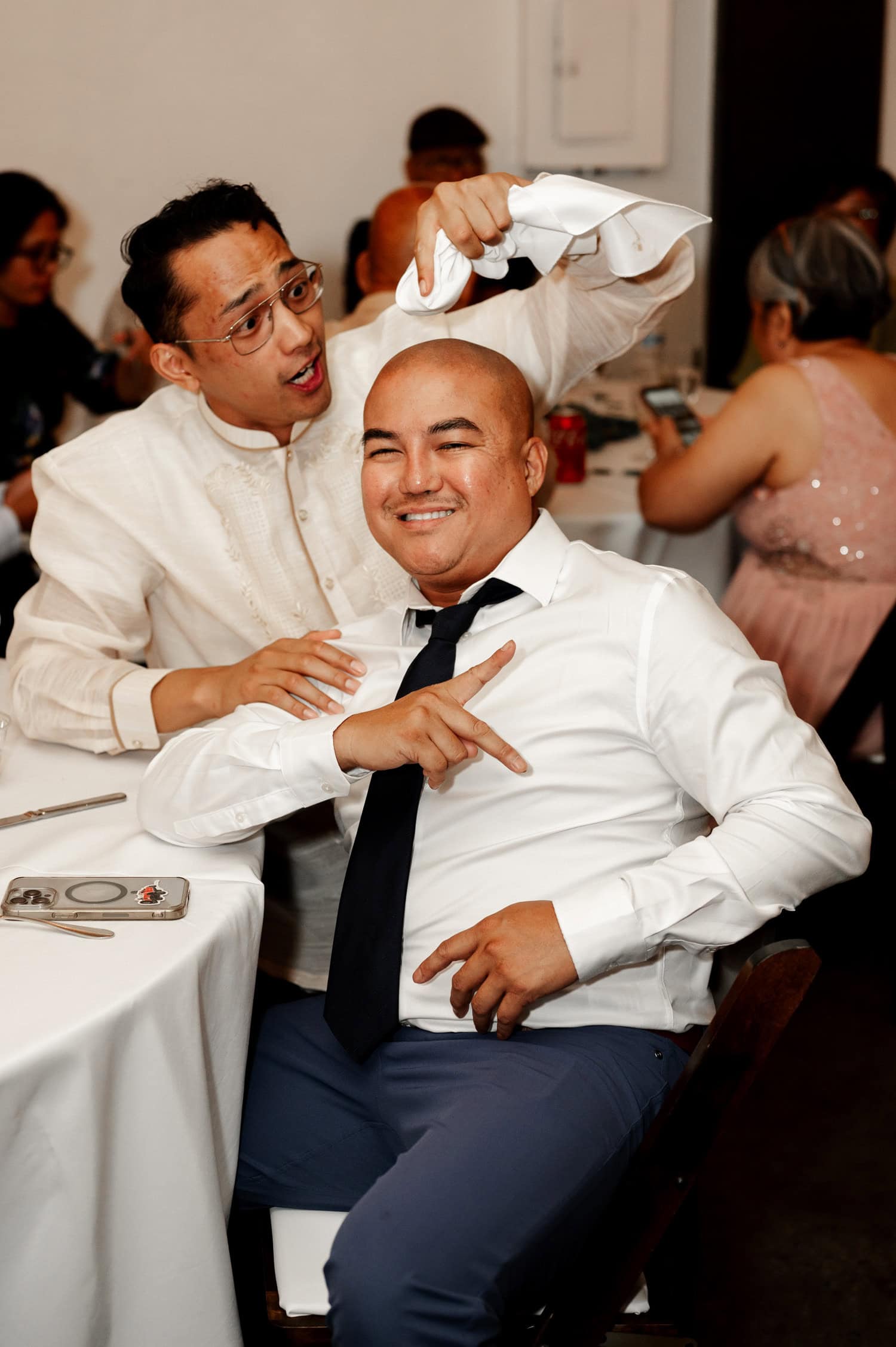 At a lively wedding at Union/Pine, a man joyfully holds a napkin above his smiling friends head as they both laugh.